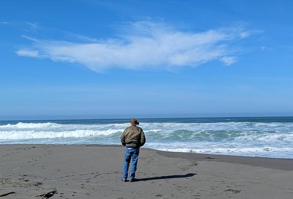 An older man wearing blue jeans and a green jacket and a leather hat is standing on a Pacific coast beach.