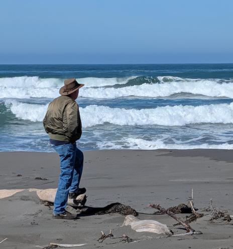 An older man wearing blue jeans and a green jacket and a leather hat is standing on a Pacific coast beach. His left foot is on a driftwood log.