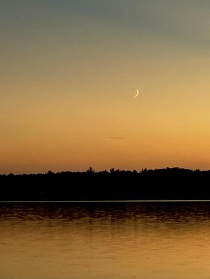 Sliver of a crescent moon in brownish orange skies over a lake after sunset