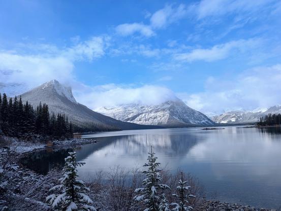 mountain scenery in the background and a lake in the foreground, there is snow on the trees.