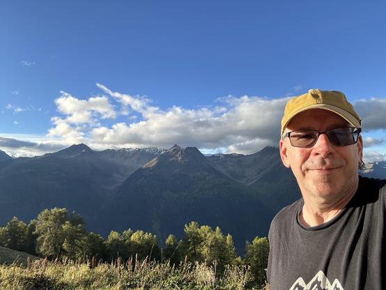 Selfie of me, a middle-aged white guy in sunglasses, t-shirt, and a mustard ball cap, in front of the view from Alp Sura (2,118 m) with Piz Giarsinom in the center.