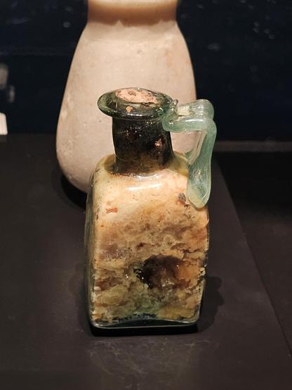 A small Roman square glass bottle with a short neck and a rounded rim, featuring a applied handle on one side. The translucent greenish glass is weathered, and the bottle is filled with pale yellow-brown residue. The vessel sits on a dark display surface, with another stone container blurred in the background.