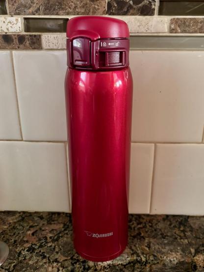 A tall narrow bright red thermos sitting on a marble kitchen counter against a tiled wall. At the bottom is the make of the thermos: Zojirushi