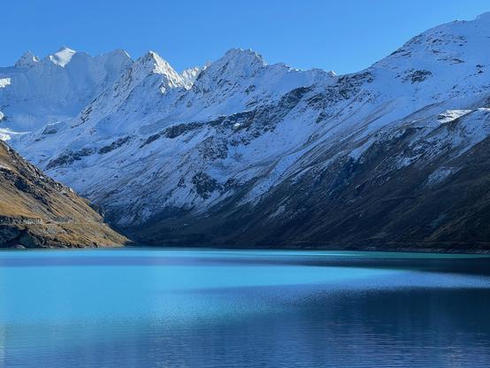 Lac de Moiry above Grimentz: the turquoise water of the reservoir fills the foreground, rippled and partly in shadow. Behind it, a long ridge of jagged peaks and steep slopes is freshly covered in snow under a clear blue sky, while the lower slopes on either side remain brown and autumnal.