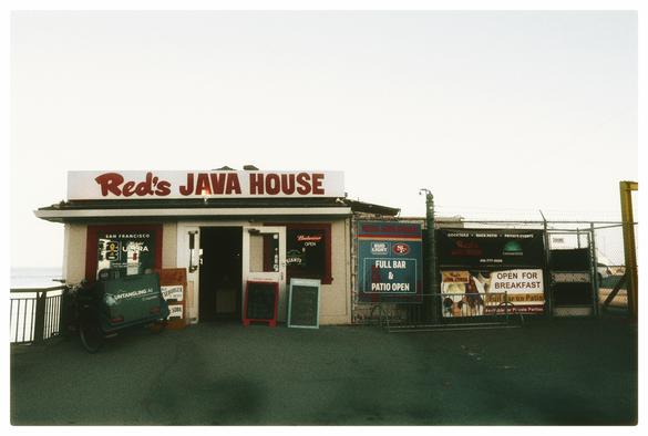 A picture of a small, single story, greasy spoon restaurant on the San Francisco waterfront. Photographed under foggy skies and looking like a shack with a chain link fence and lots of signage on a stretch of asphalt tarmac, Red's Java House is a never-changing San Francisco institution, famous for basic ballpark food, fast service, beers and no-nonsense Bloody Mary's.