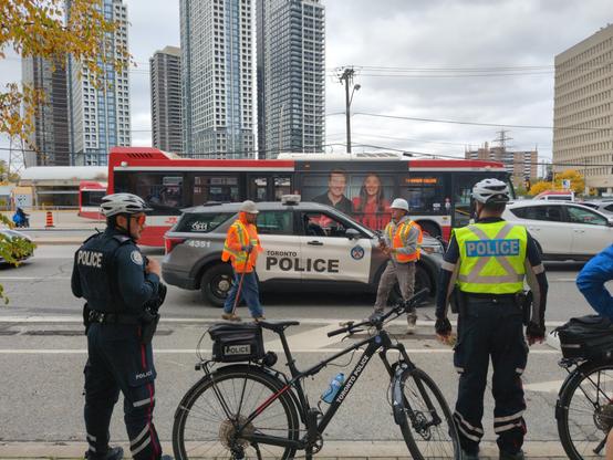 On the curb, two cops stand guard as two construction workers stand near the patches where concrete curb things used to be. A TTC bus coming from Islington station is in the background.