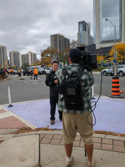 A rather meta shot of a TV cameraperson filming a clean-cut guy with a microphone, while bike lanes are removed in the background.