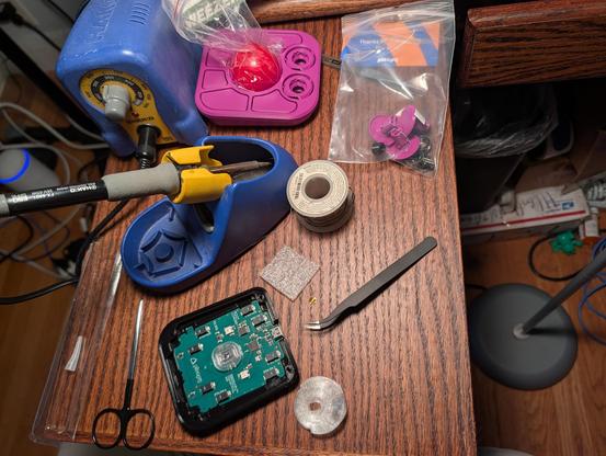 In the bottom of the image, a printed circuit board. Above that, a yellow and blue soldering iron that looks like a child's toy. Above that, a purple trackball top with a bright red ball sitting inside the hollow of the case.