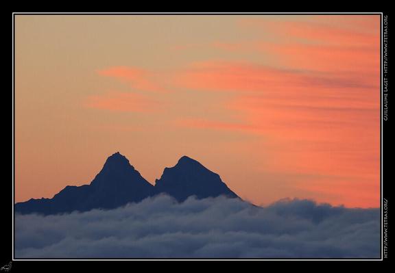 Deux sommets pointus émergent d'une mer de nuages, dans un ciel rougeâtre avant le lever de soleil