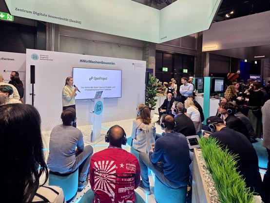A presentation at a digital conference showcasing OpenProject, with a speaker addressing the audience. Attendees, some wearing headphones, are seated and engaged, while others stand nearby. The setting includes modern decor and branding related to digital sovereignty.