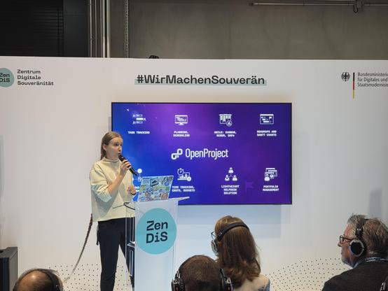 A woman speaks at a podium during a presentation at an event, with a large screen behind her displaying information about project management tools, including task tracking, planning, and scheduling. Attendees wearing headphones are seated in front of her.