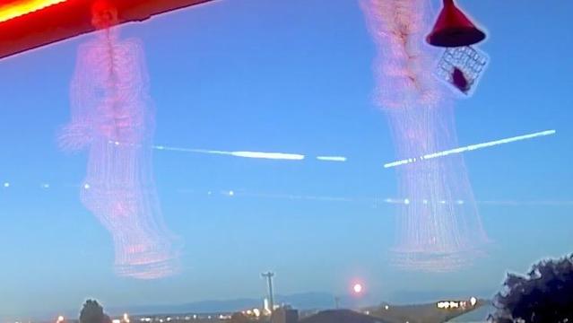 A view of the sky at dusk with faint silhouettes of skeleton figures, illuminated by red lights. A bird feeder is visible in the upper right corner, and a distant horizon features hills and lights. Streaks cross the sky, the bottom and right are from planes and top-middle one is from space debris burning up. This is an image generated by stacking video frames.