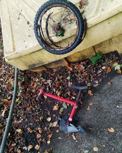 The remains of a red child's bicycle. 
The foreground is grey tarmac and earth, with fallen autumn leave, mostly beech. The background is a yellow salt-grit bin, upon which is perched a rusted out wheel and tyre.
All that's left of the bike is the top tube, a bit of headstock and part of the seat tube & post, and the black saddle.