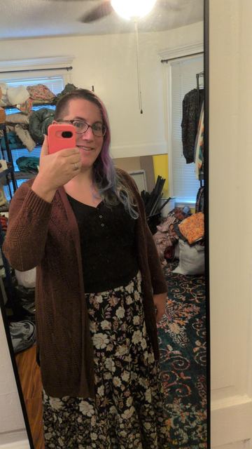Mirror selfie of a smiling woman with glasses and long, multicolored purple, blue, and green hair, shaved on one side. She wears a black button-front top, a long brown knit cardigan, and a black maxi skirt with a cream and pastel floral print.