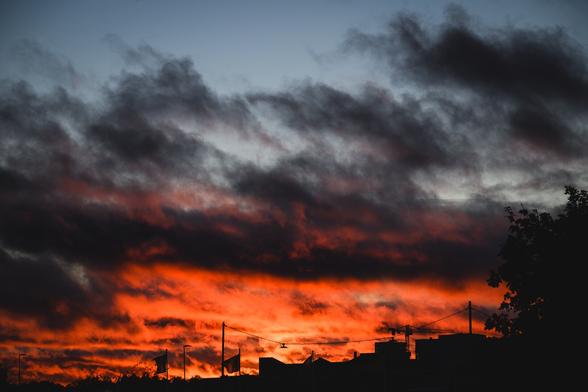 A photograph of an intense sunset. In the silhouetted foreground are the tops of a few buildings along the bottom edge with a tree in the lower right corner.

The lower half of the photograph is dominated by an intense orange sunset, illuminating the half cloudy sky. Where the clouds are not reached by the formidable sunlight, they loom dark and grey.

Along the top edge the sky clears up with a band of clear and quite bright blue.