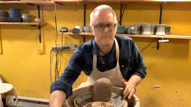 A time-lapse video of a middle-aged man with short white hair, glasses, a blue plaid shirt, and a white apron working at a pottery wheel in a studio with bright yellow walls and wooden shelves holding pottery. The footage is sped up as he quickly centers the clay, shapes a tall cylinder, and then opens and flares it outward into a wide bowl. There is no sound.