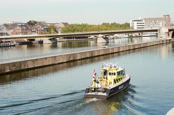 A yellow and black boat with a red flag sails on a river, with a bridge and buildings in the background. The text on the boat reads "N6326".