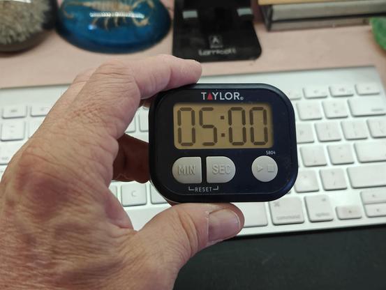 A cheap LCD digital kitchen timer, held in a human hand. There is a computer keyboard and desk clutter in the background.