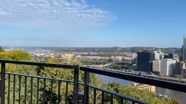 Video taken from a high hill overlooking Pittsburgh from the Monongahela side. It’s a beautiful blue sky day and the city looks beautiful in the sunlight, against the blue sky. Out past the city are more wooded hills with some buildings on them.