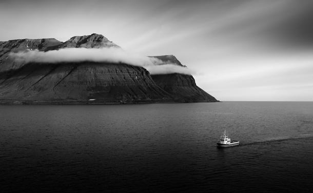 This is a monochrome image of a boat, mountains and clouds in Isafjordur, in Iceland. The thin ribbon of clouds wrap themselves around the top of the mountains, giving them a feeling of being some kind of headband. The small boat, heading towards the viewer, pulls the eye down to the corner of the image.