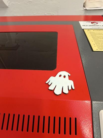 A small white spooky ghost sits on top of the red lid of a lasercutter.
