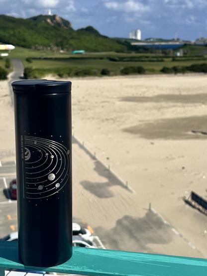 A flask with a design showing the solar system sits in a ledge overlooking a beach. In the background, out of focus, is the launch pad.