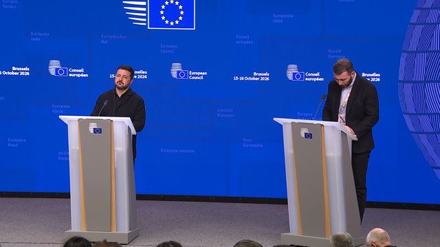 A journalist stands in a crowded press room holding a microphone and asks a question. Cut to Volodymyr Zelensky at a podium in front of a blue European Council backdrop reading "Bruxelles 15-16 octobre 2026". He looks down in silence for about ten seconds, then looks up and says: "This is my answer. Thank you so much." Smiling, he takes out his interpretation earpiece and turns away from the podium.