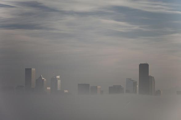 A city skyline partially obscured by fog, with several tall buildings and a cloudy sky in the background. The scene conveys a moody atmosphere.