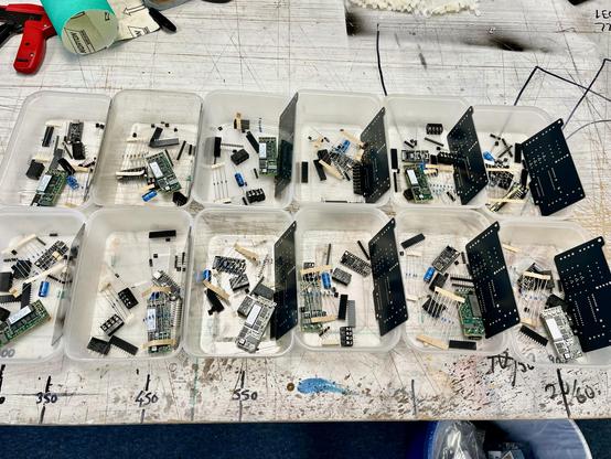 12 plastic takeaway trays arranged on the studio bench, each filled with an identical set of through-hole components, small daughterboards and a bare black PCB.