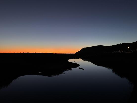 disk over a tidal lagoon. a thin ribbon of orange stripes the horizon with still dark water in the foreground and silhouetted headlands