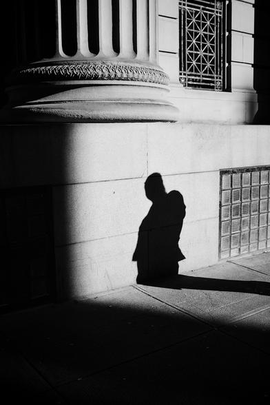 Outside the impressive Bank of Scotland building in Glasgow city centre, where the autumnal sunlight is creating patches of light and dark shadows on the towering stone columns and the shadow of a person approaching on the street bends toward the foot of one of the columns. The person is out of frame.