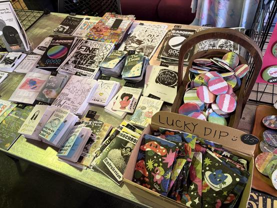 Zines : small brochure like publications. Spread on table next to some lucky dips and queer “flag” badges in a timber basket.