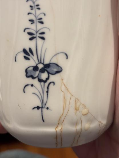 Close-up of a white fluted porcelain vessel with a hand-painted cobalt blue floral sprig. Near the base, a hairline crack has absorbed tea, leaving thin amber stain lines that branch and trickle down toward the bottom edge, along with a few small cream-colored drips.