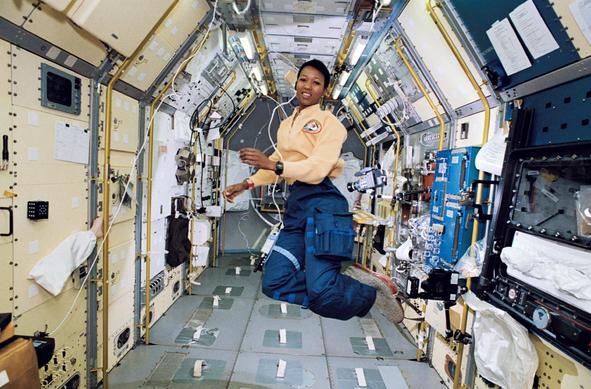 Black woman floating in a corridor of the international space station. She is wearing a yellow blouse and blue pants.