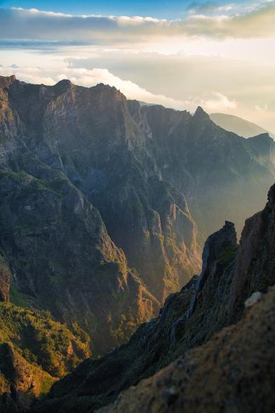 High perspective view into a deep mountain gorge; steep slopes covered in green vegetation are bathed in warm golden hour sunlight through cool mist.