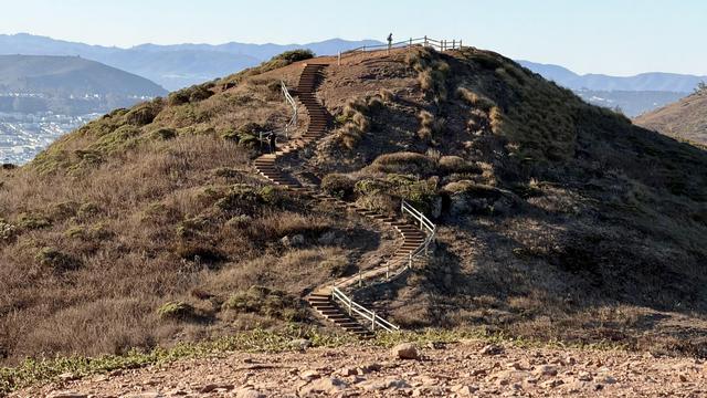 A person stands on a hill with a staircase leading up to a railing. The hill is covered in dry grass and bushes, with a city visible in the distance.