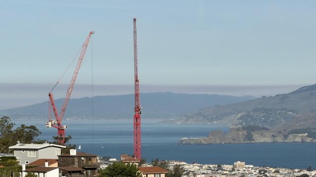 Two red cranes overlooking a city and a body of water under a clear blue sky.