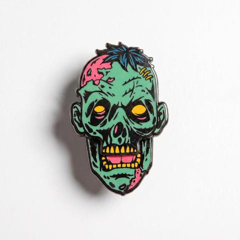 Enamel pin depicting a cartoon green zombie head with glowing yellow eyes, pink brain exposed, yellow stitches, and spiky dark hair on a plain white background.