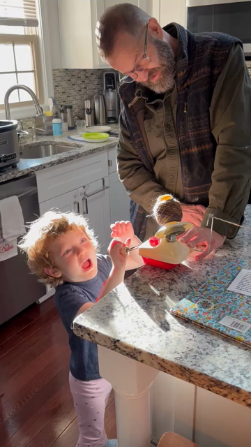 An adult and a young child with curly, light-colored hair stand at a speckled granite kitchen island, winding yarn onto a small tabletop ball winder. The child, wearing a dark T-shirt and light patterned pants, looks up intently while helping turn the handle, with sunlight streaming across them from a window.

Sound description: A cozy kitchen ambiance featuring the steady, rhythmic whir and click of a mechanical yarn winder, accompanied by the gentle, conversational voices of an adult and a young child talking and encouraging one another.