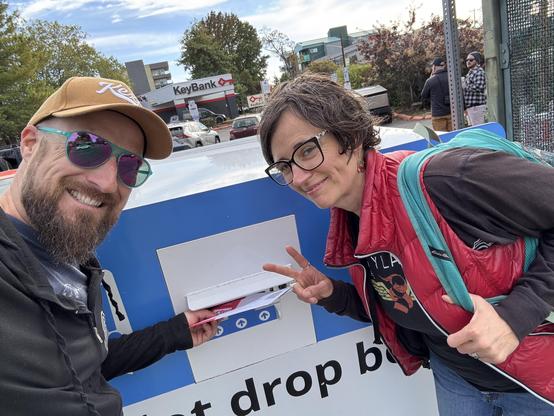 A man and a woman pose in front of a ballot collection box, both smiling. The man has a beard and wears sunglasses and a brown cap, while the woman flashes a peace sign. In the background, a KeyBank and other buildings are visible