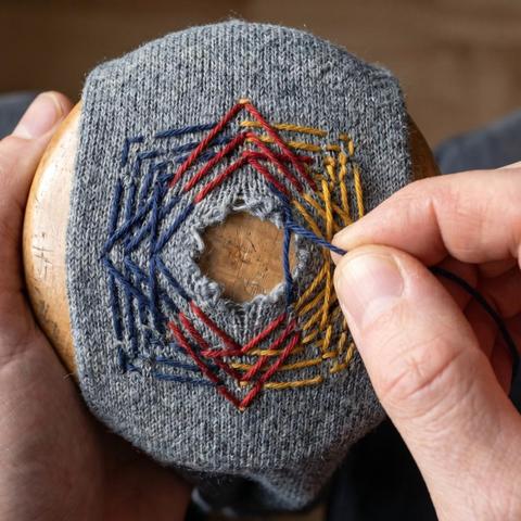 Hands darning a hole in a gray knitted sock with colorful red, blue, and yellow threads stretched over a wooden darning egg.
