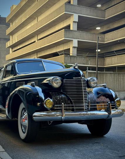 An antique black car with an intricate chrome front grill, bumper, and lights. Everything is round, streamlined. A plaque on the right says: 1940 World’s Fair New York, New York. 
Most likely not silent when motoring, but here it is in a back downtown street, silent.