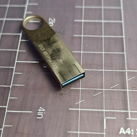 A slim metal USB stick on a cutting mat. You can see that the metal at the "port" end is a bit bent and scratched. It says 64GB, but it's barely readable