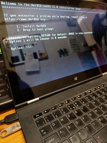 Thin laptop with a shiny screen showing the NetBSD loader. Option 1, Install NetBSD, is about to be selected. Reflected int ehbsxreen you can see a fairly interesting part of the office wall with pictures and whiteboard on it. Slightly blurry.