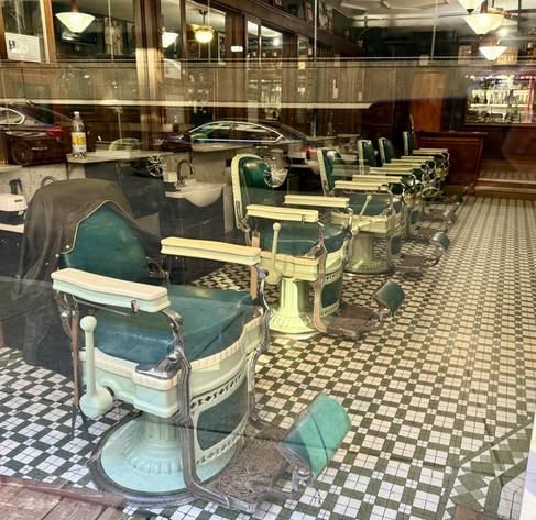 View through a window into the closed Patsy's Barber Shop in Albany, NY. There's a line of six intricate metal barber shop chairs anchored into a black and white tiled floor. Each chair has a large handle to lift/lower the patron. The foot and arm rests are all hinges so they can be tilted back for hair washing. Seats are a dark green, possibly leather covered cushions. The window we are looking through is reflecting a couple modern cars from out on the street, juxtaposition between their metal and plastic lines and the old, solid materials making up the shop within.