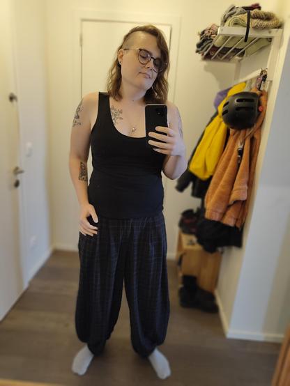 Mirror selfie of me in a black tank top and sort of poofy tartan pants, I look chill and cute.