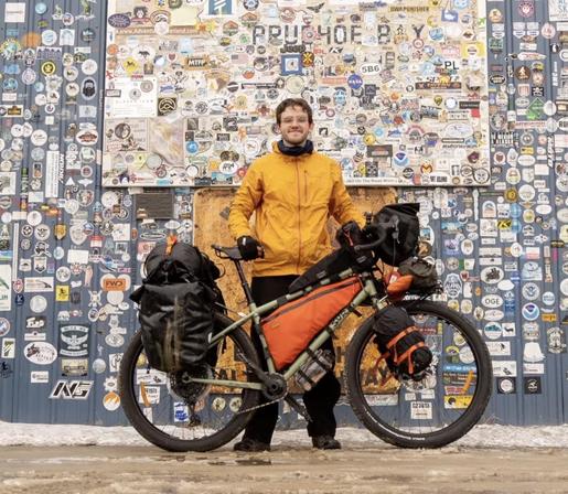 Dan 3 years ago and Prudhoe Bay Alaska posing with his very loaded bikepacking setup in front of a wall full of stickers, from previous travelers.  His bike is light green, what you can see of it, with a large orange frame bag, large panniers on the back, round stuff sacks on the front forks, a handlebar bag, another bag below that on small rack, top tube bag, water bottle under frame in front of bottom bracking, and another large pack on top of the back rack. The bike is a 1x, probably 1x11.