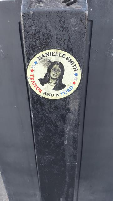 A round sticker of Daniel Smith's face with the words "DANIEL SMITH TRAITOR AND A TURD".
The sticker was on an outdoor cigarette ashtray stand and shows marks indicating that people have stubbed out their cigarettes on the image of her face.