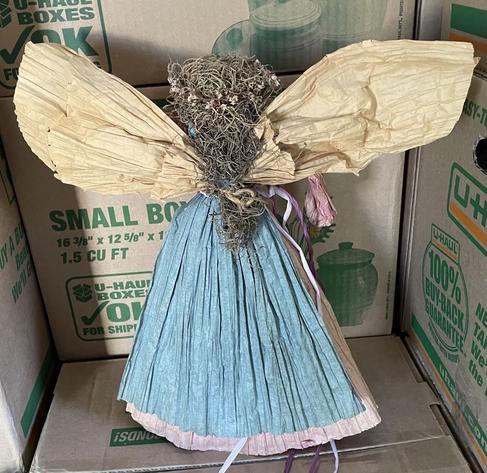 From the back the figure’s skirt is blue. Her dark hair tumbles down her back between pale gold colored wings.
