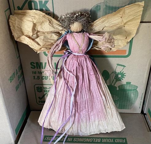 A doll hand-fashioned from paper. Its paper dress is pink ombré and its wings are a golden beige, the color of ripe wheat. On its hair is a crown of tiny dried flowers.